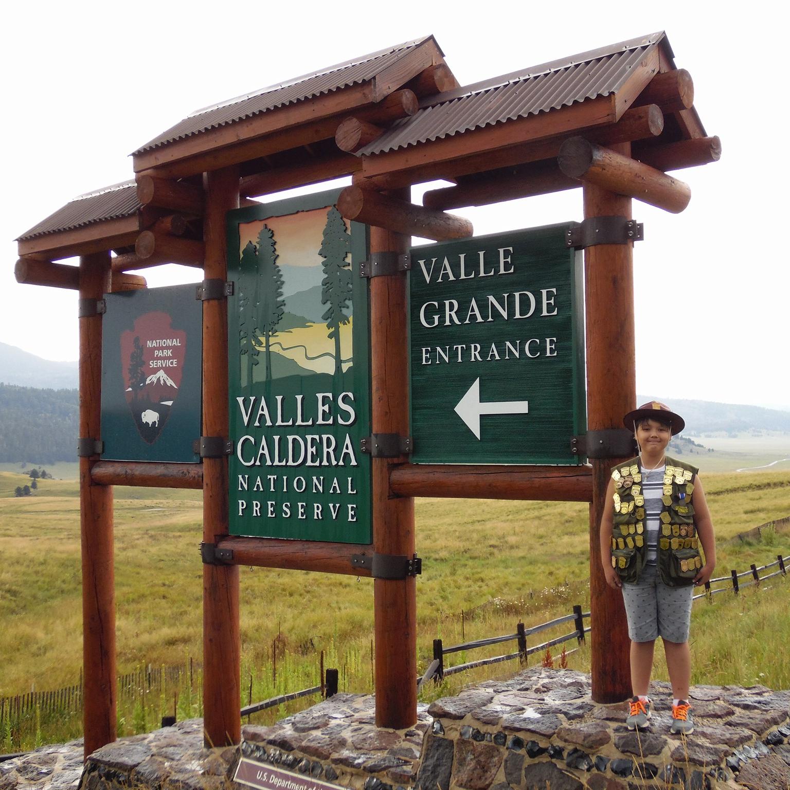 A boy wearing a Junior Ranger hat and vest filled with badges stands at Valles Caldera\'s sign.