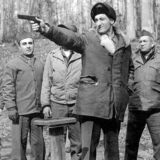 a group of men standing in a wooded rifle range, in foreground one has pistol raised