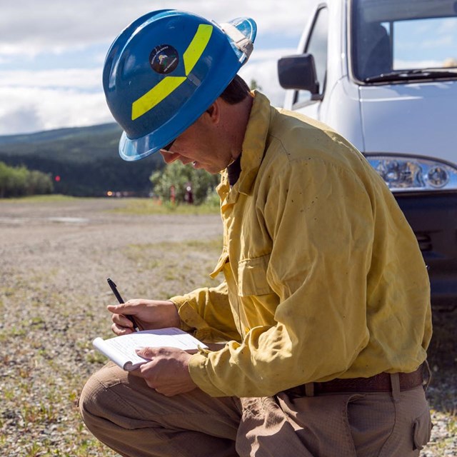 A wildland fire crew member kneels down in gravel to fill out his time sheet.