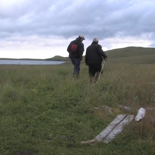two people walk through a grassy field with old lumber and the ocean in the distance.