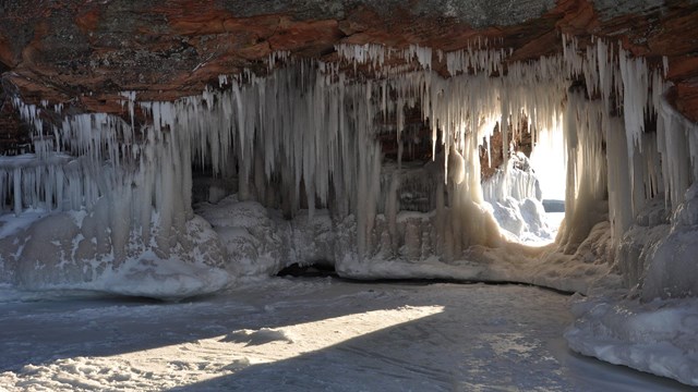 Photograph inside a sandstone cave covered in ice with sunlight streaming in through an opening.