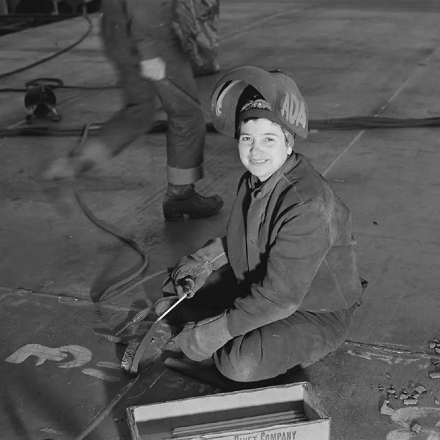 A woman kneels on the floor in a welders uniform, she is smiling at the camera.