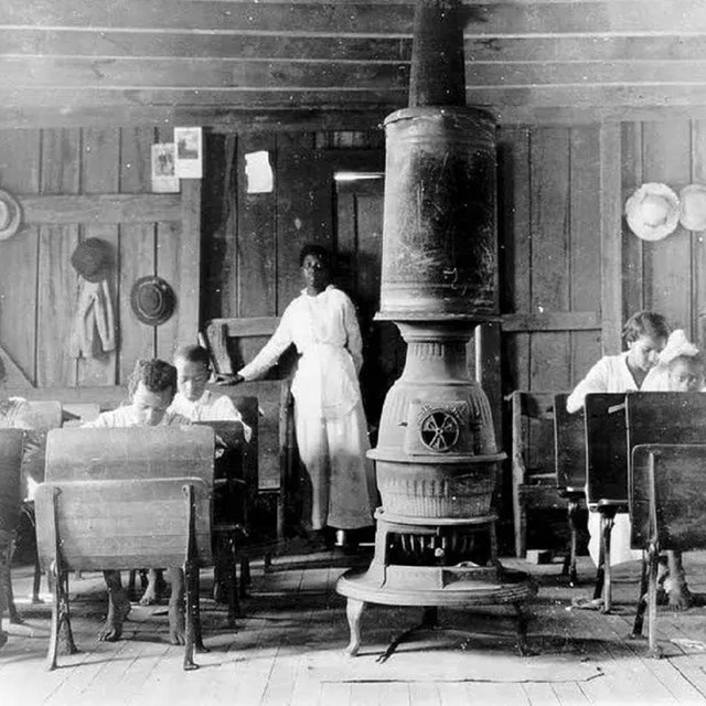 Black and white image of children sitting in a classroom with a stove in the center