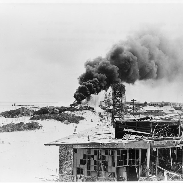 black and white photo of beach with destroyed building in foreground and smoke in background