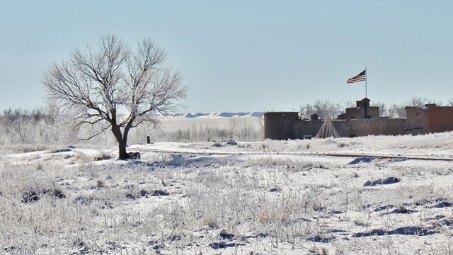 A snow covered landscape with the tall walls of the adobe fort in the foreground. 