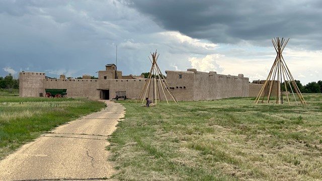A paved walkway leads to an adobe fortress with tipi poles in front of it