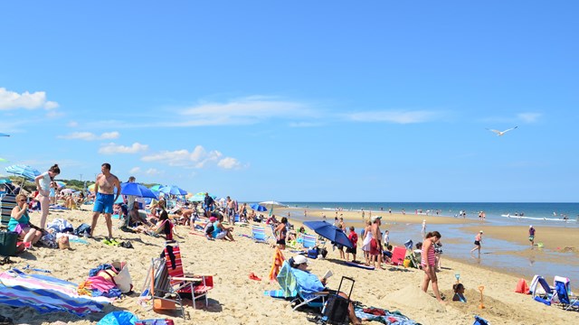 A crowd with colorful blankets and umbrellas enjoys a a sunny beach day.