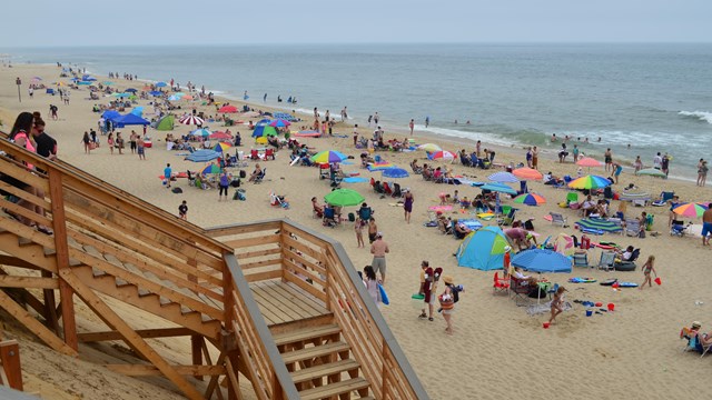 Wooden stairs lead down to a beach with many multi-colored blankets and umbrellas.