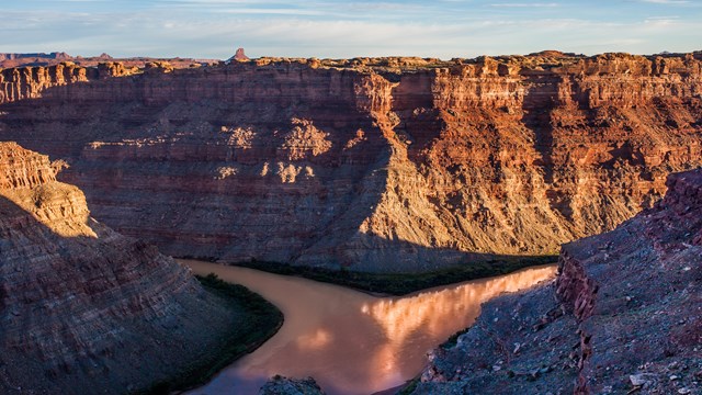 Two rivers converge at sunset against the backdrop of a towering, orange and brown canyon wall. 