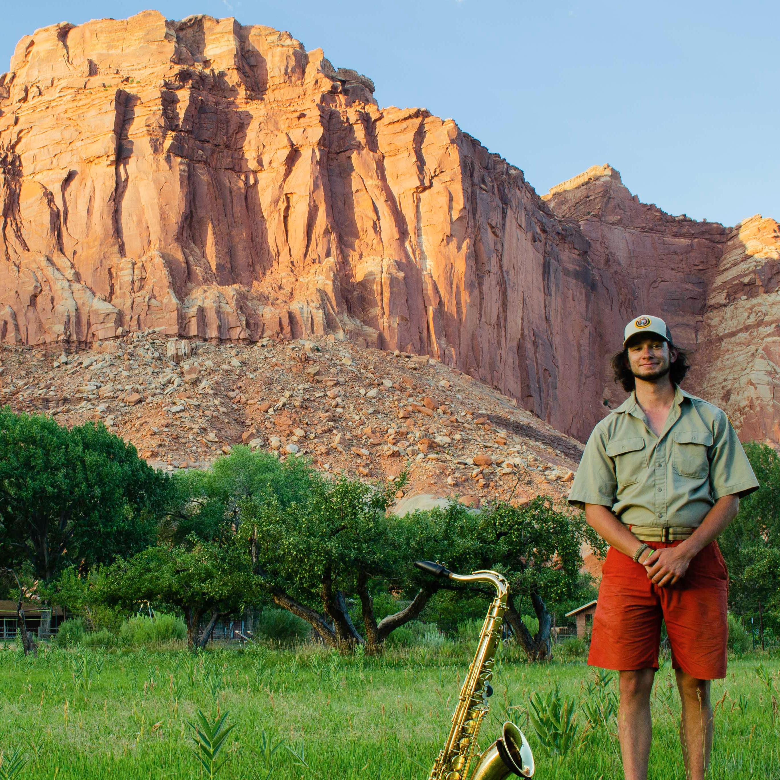 A man standing in a green field next to a gold saxophone with orange rock cliffs in background.