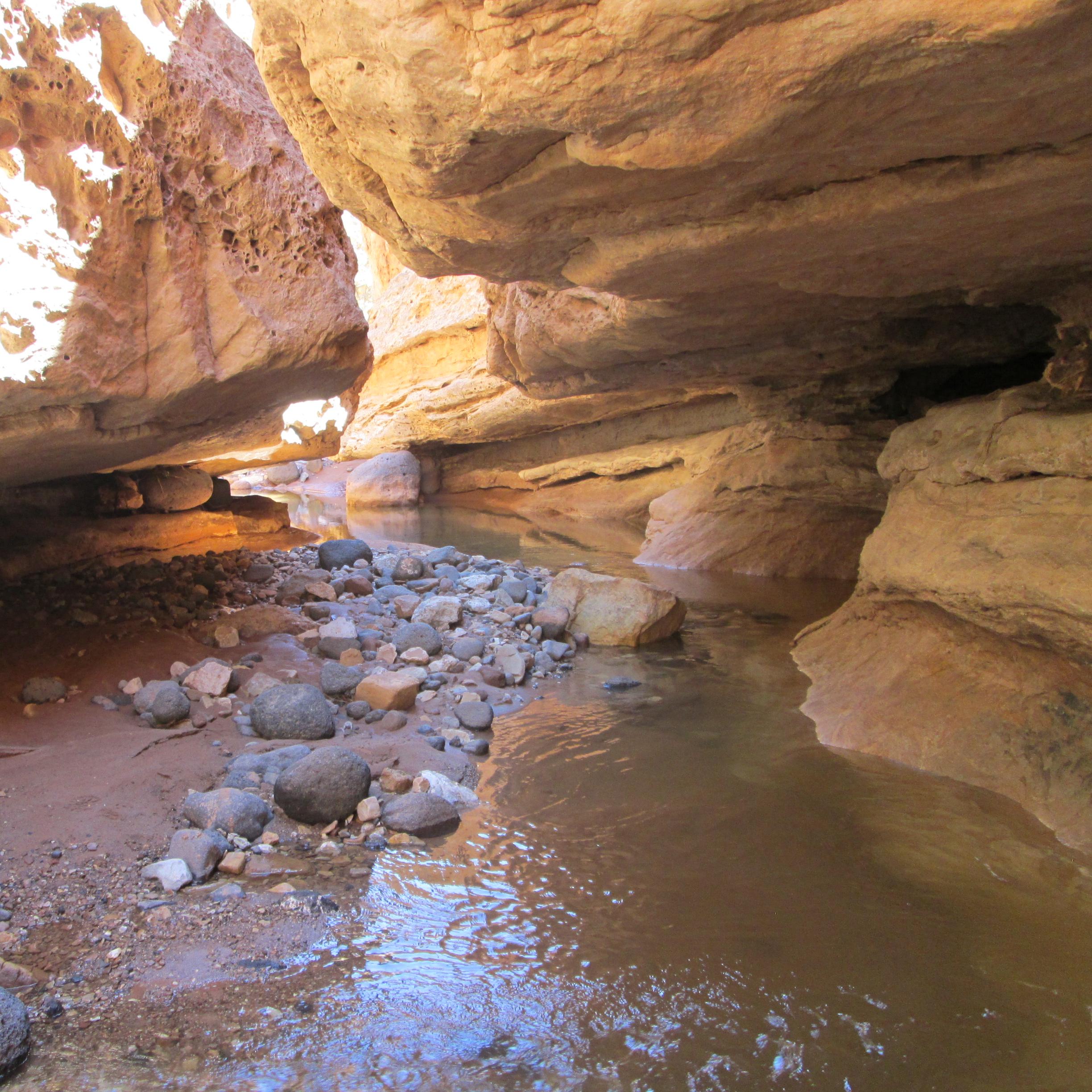 Canyon with overhanging rock walls, water, and black boulders.