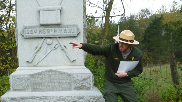 A park ranger next to a war memorial points emphatically. 