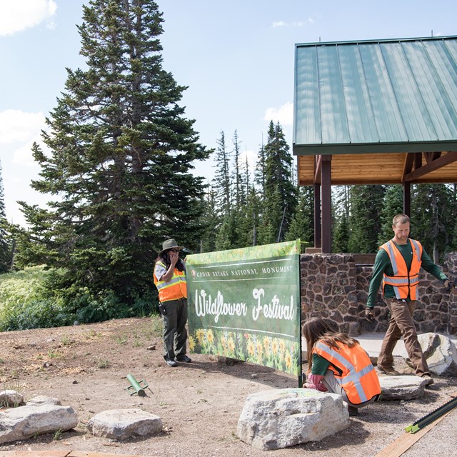 Three park employees install a special event sign in front of a pavilion