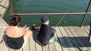 Two people sit on a dock, holding fishing rods.
