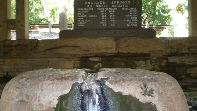 Water flows up out of a rock. An old wood routed sign details the mineral content of the water.