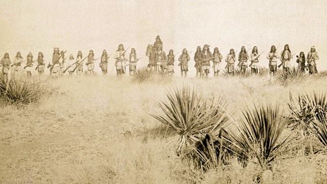 Sepia-toned photograph of one man on a horse, and a line of warriors on foot. 