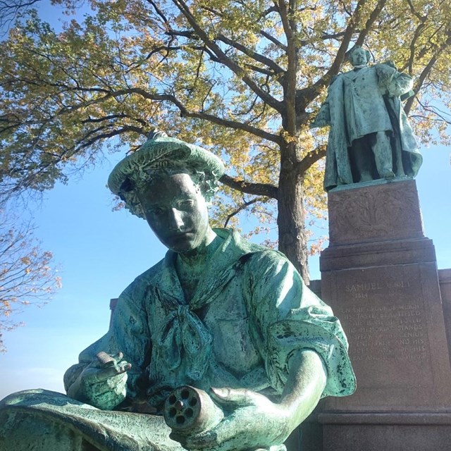 A blue/green monument with two figures: a boy sitting and carving and a man standing tall. 