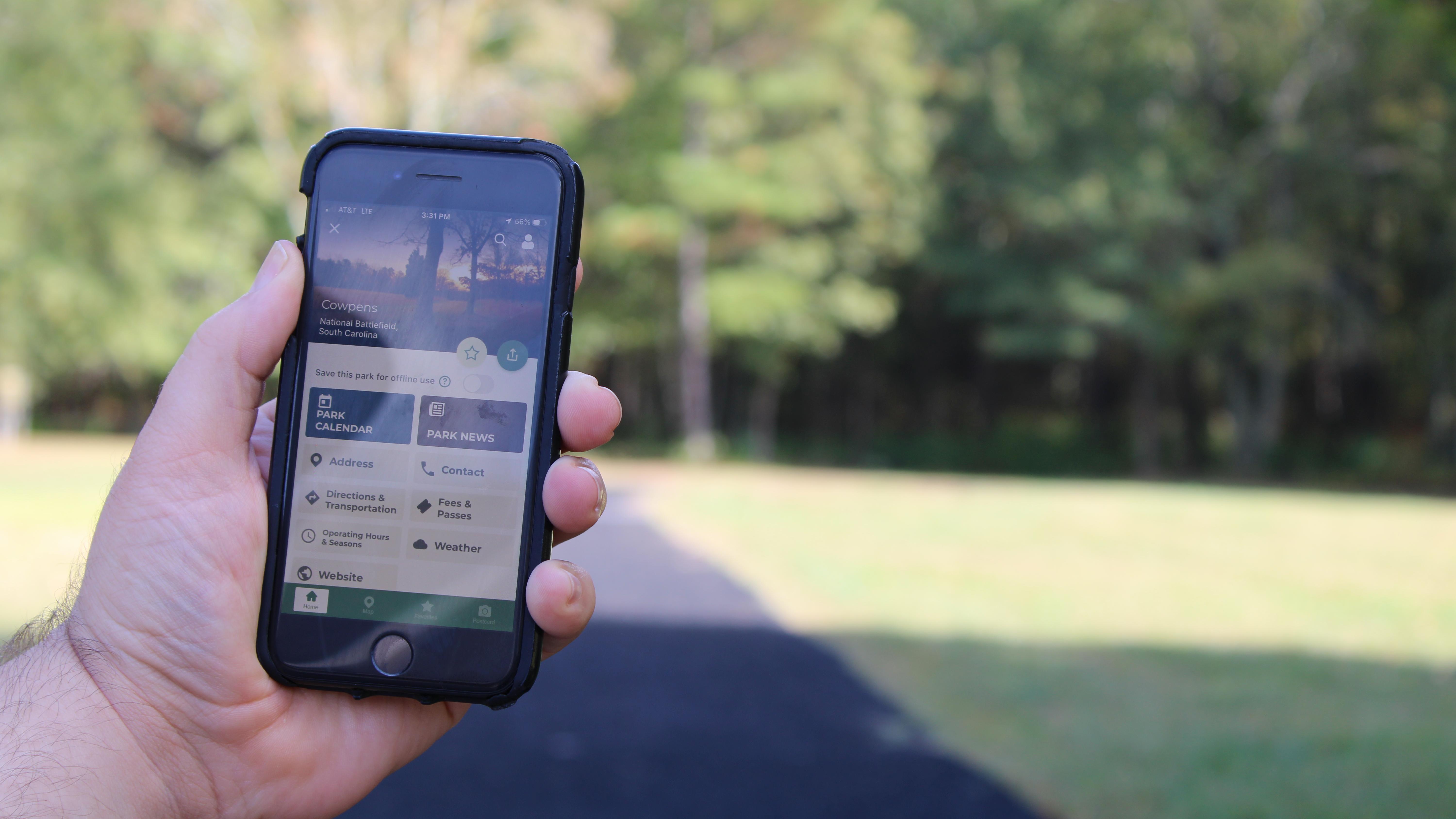 Hand holds cell phone with mobile app. Trail is seen in the background. 