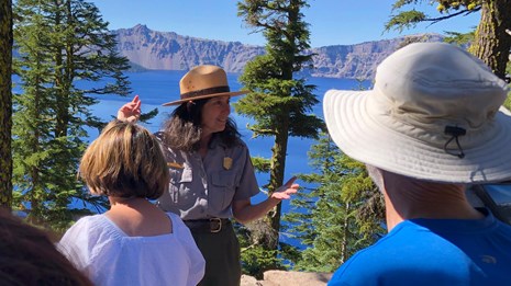 Female Ranger talks with a group of visitors outside with conifers and Crater Lake behind her.