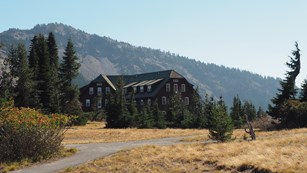 A side view of Crater Lake Lodge, forest, grassy meadow and Garfield Peak rising behind it.
