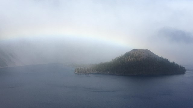 Mostly cloudy lake view with a rainbow from the caldera rim to the top of Wizard Island, a volcano. 