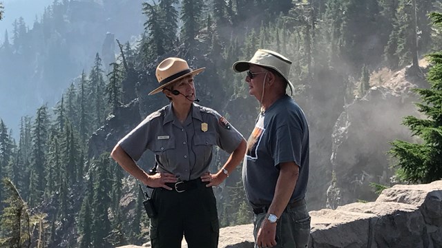 A ranger talks with a man with a stone wall and forest behind them. 