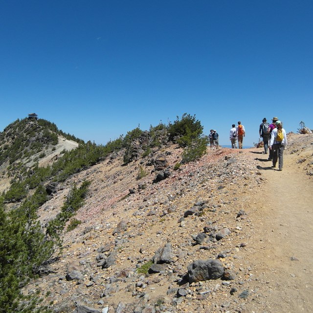Hikers on a high elevation trail with sparce vegetation to Mount Scott Fire Lookout in the distance