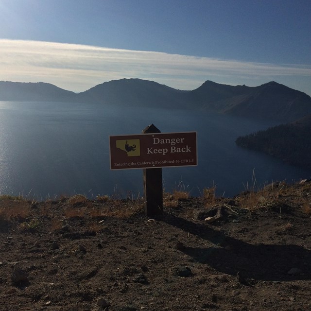 A wood sign with Danger painted in white is at the caldera edge with lake behind. 