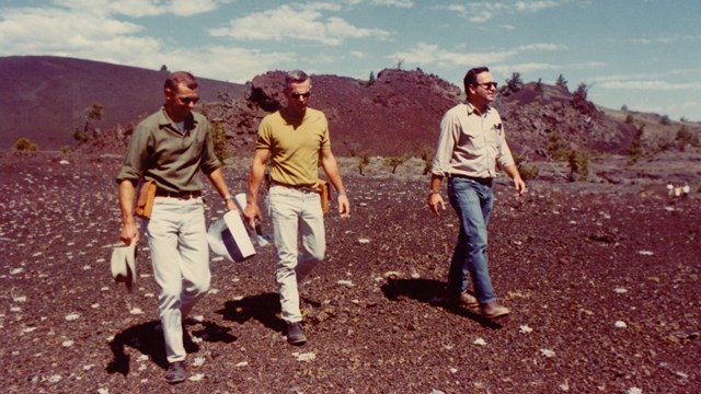 three men walking across a black, rocky landscape with the text "NASA" in the corner