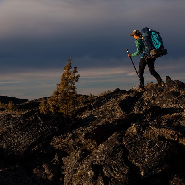 Woman with backpack and hiking poles traverses a rocky, lava landscape. 