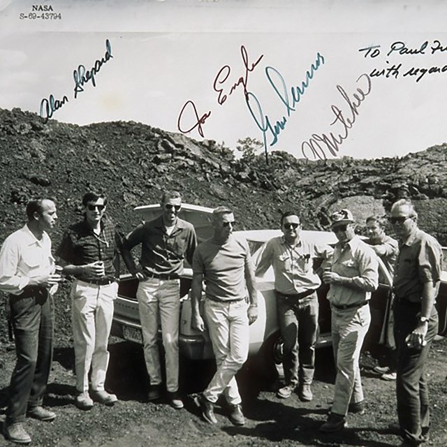 Eight men stand in front of a car parked in a lava landscape. 