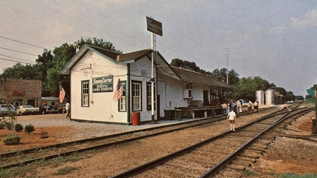 A photo postcard from the late 1970s shows people near railroad tracks to the right of a depot 