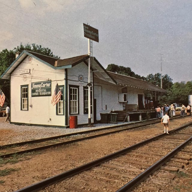 A photo postcard shows people near railroad tracks to the right of a depot in Plains, GA, late 1970s