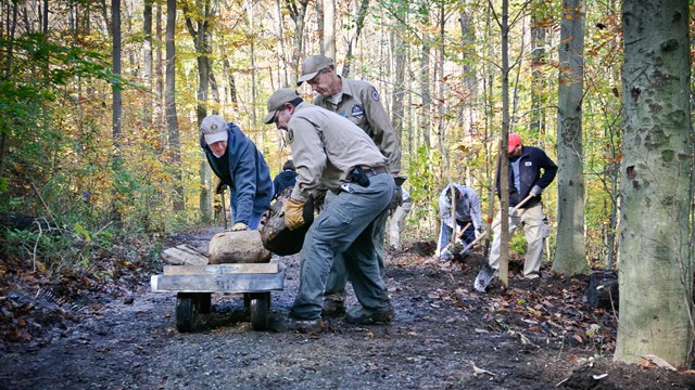 Three men in khaki shirts and hats lift a boulder onto a cart on a wooded trail.