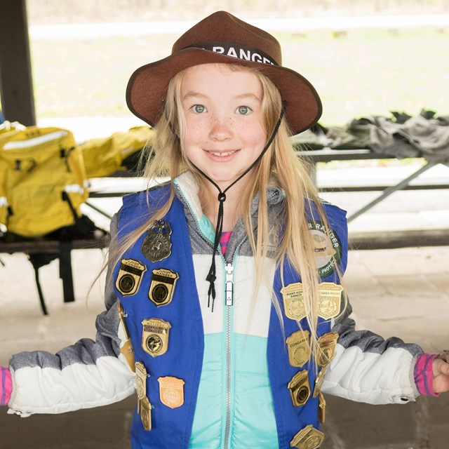 A girl wearing a brown Junior Ranger hat and a blue vest covered in badges smiles at the camera