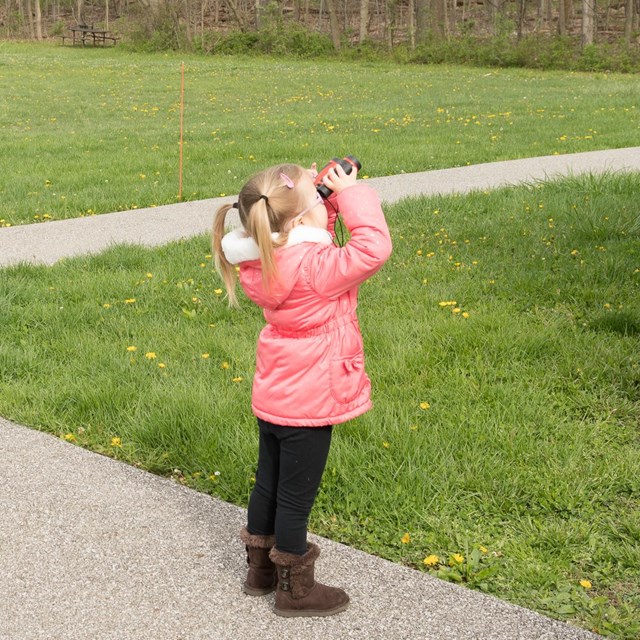 A young girl in a pink coat standing on a path looks off to the right through a pair of binoculars