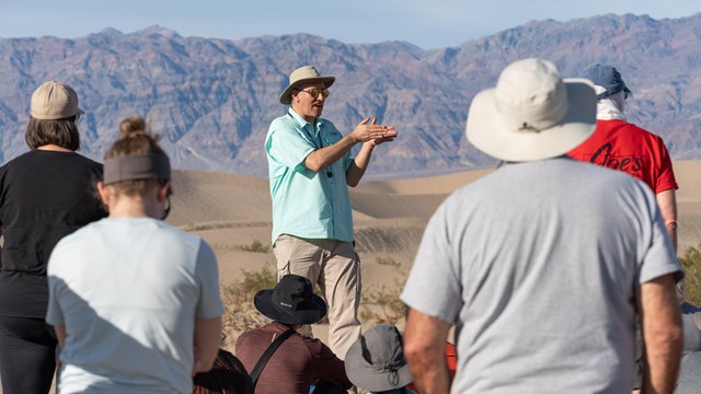 A man standing on sand dunes talks to a crowd with mountains in the background.