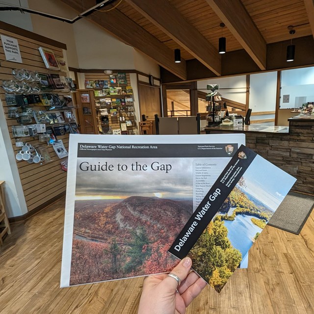 A hand holding out a guide and a park map with the visitor center in the background.