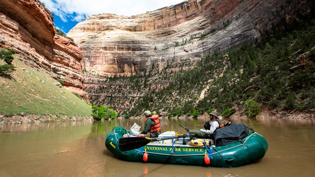 Rafters clad in life jackets float down a river in a green National Park Service raft.