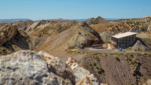 Peering over a rocky outcrop, the camera looks down at a building built into a colorful hillside.