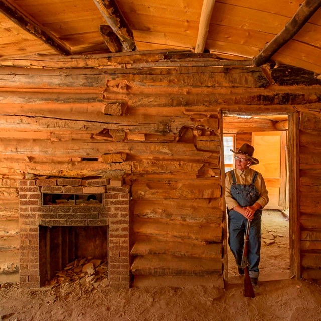 A living history actress stands in the doorway inside of a log cabin next to a brick fireplace. 