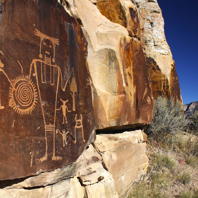 An image of a large humanoid figure with a shield and smaller figures etched into a rock face.