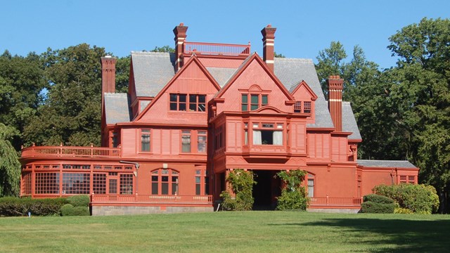 A three story brick red mansion on a lawn surrounded by trees.