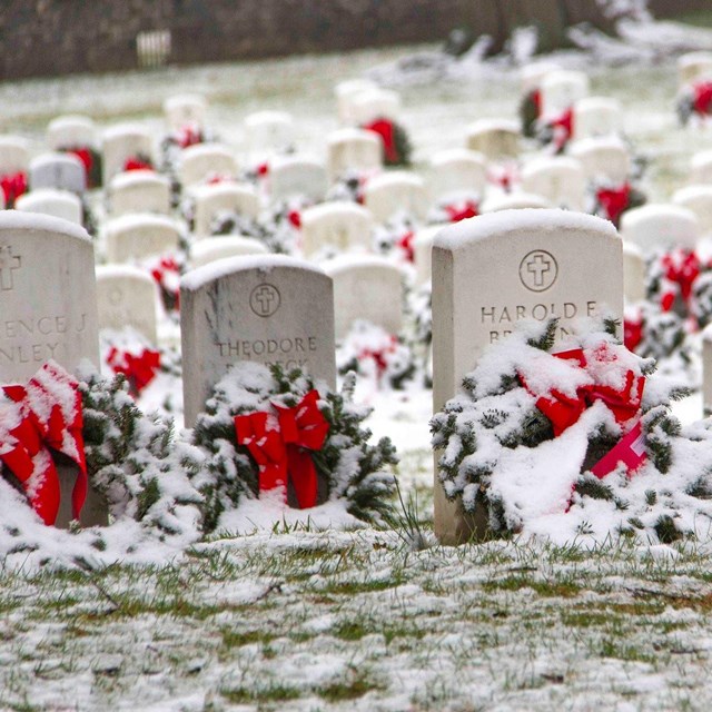 A color image of wreaths and red bows on white headstones covered in snow