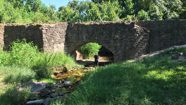 A person stands underneath a stone arch in a vegetated area.