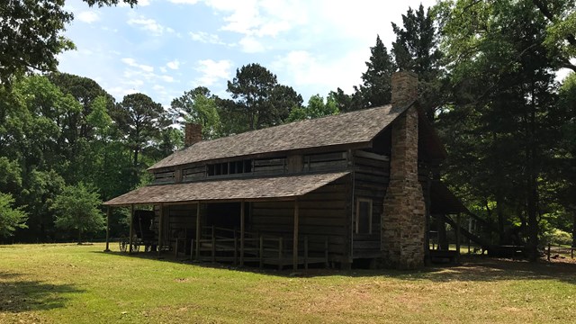 A wooden long house set on a grassy field, surrounded by trees.