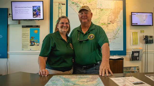 Two Park Volunteers behind a desk at the Visitor Center pose and smile.