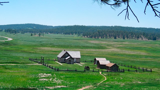 Four old log buildings and wagon surrounded by a wooden fence in a green field under a blue sky.