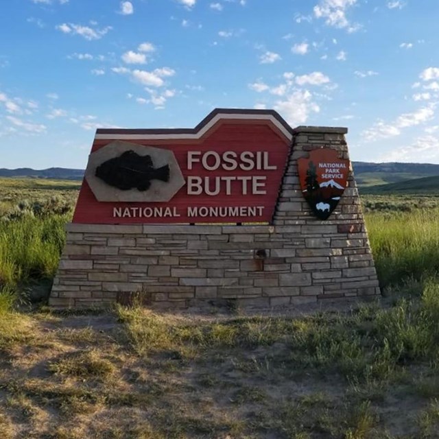 Sign that says Fossil Butte National Monument with fossil fish and arrowhead. Sun rising over butte.