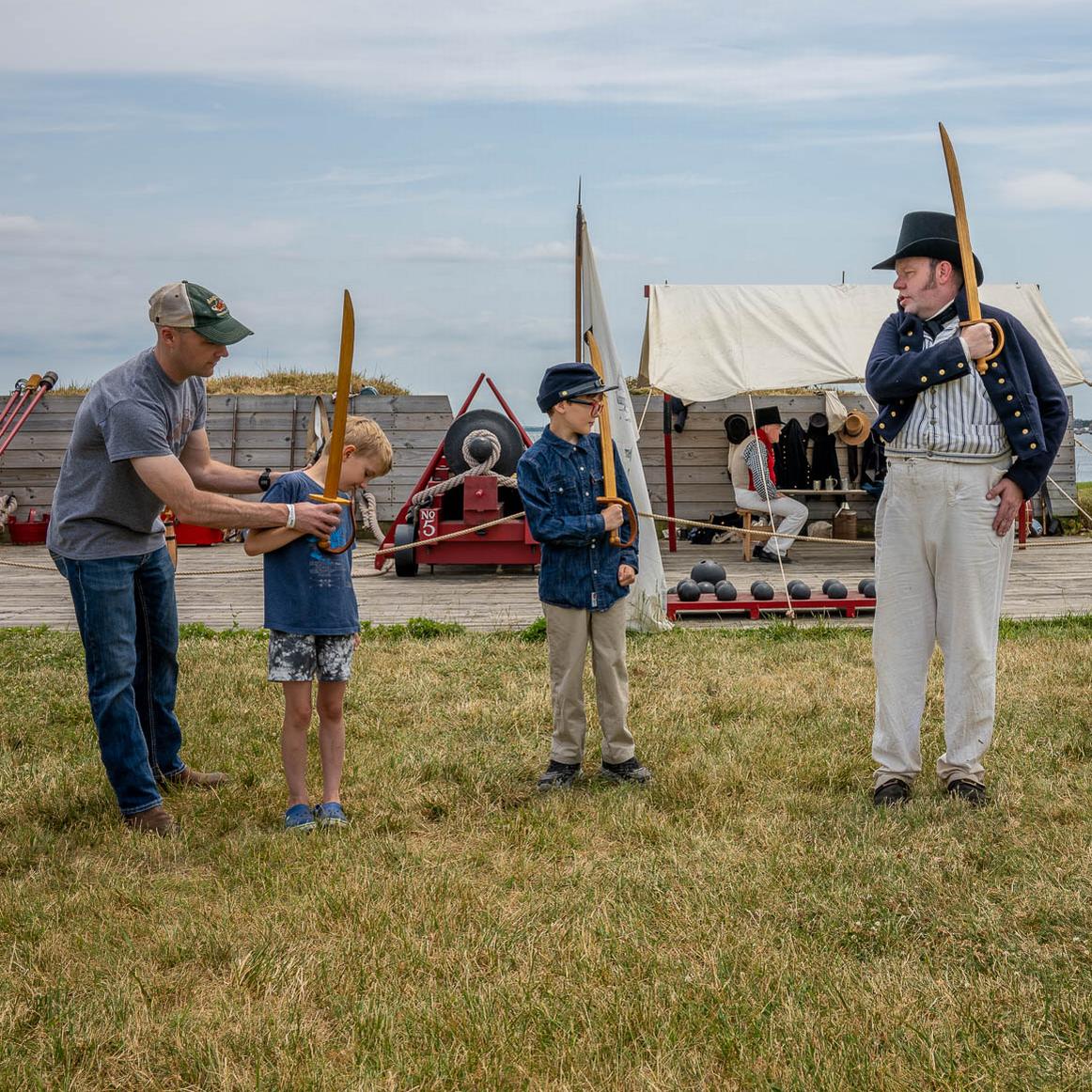 A living history member instructing two children and one adult during a wooden cutlass drill.